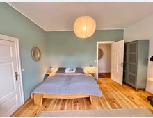 Ein Schlafzimmer mit Holzfußboden, einem Doppelbett mit grauer und rosa Bettwäsche, und einer runden Deckenleuchte. Wände sind grün gestrichen, rechts steht ein grauer Schrank, und links sowie im Hintergrund sind weiße Türen sichtbar.