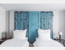 Schlafzimmer mit zwei Einzelbetten und weißer Bettwäsche, eingerahmt von einem rustikalen blauen Holzpaneel als dekoratives Kopfteil. Auf den Nachttischen stehen eine kleine Lampe und ein dekorativer Vogelkäfig.