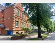 Zweistöckiges Wohnhaus im historischem Stil mit orangefarbener Fassade, hellen Fensterumrahmungen und einem spitzen Giebel. Davor ein gepflegter Garten mit Metallzaun, einer mit Pflastersteinen belegten Gehweg- und Straßenfläche sowie großen Bäumen am Straßenrand.