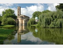 Historisches Kirchengebäude mit einem rechteckigen Glockenturm im romanischen Stil, umgeben von grünen Bäumen und einem Teich, der die Kirche und das grüne Umfeld spiegelt; im Hintergrund ist eine Kuppelbau-Struktur sichtbar.