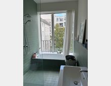 Modernes Badezimmer mit grün gefliesten Wänden und Boden, einer freistehenden Badewanne vor einem großen Fenster mit Vorhängen und Blick auf umliegende Gebäude und Bäume. Links befindet sich eine bodengleiche Dusche mit einer Glasabtrennung.