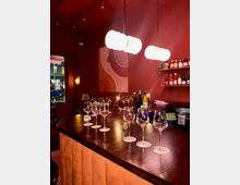 Interior of a wine bar with a long dark wooden counter set with empty wine glasses and bottles, lit by glowing pendant lights. Deep red walls feature a large abstract painting and shelves of wine bottles near a street-facing window.