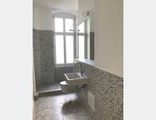 Badezimmer mit weißen Wänden, großen Fenstern und Terrazzo-Boden sowie -Wandverkleidung. Es gibt ein weißes Hängespülbecken, eine wandmontierte Toilette und eine Dusche mit runder Kopfbrause.