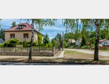 Wohngebiet mit freistehenden Einfamilienhäusern, einem gepflegten Garten und einem umzäunten Grundstück im Vordergrund. Die Straße ist von Bäumen gesäumt, im Hintergrund stehen weitere Häuser in einer grünen Umgebung.