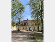 Gelbes historisches Gebäude mit roten Ziegeldächern, mehreren Dachgauben und weißen Fensterrahmen, umgeben von hohen Bäumen mit frischem Frühlingslaub. Der Vorplatz ist unbefestigt, mit sandigem Boden und einigen kleinen bepflanzten Bereichen.