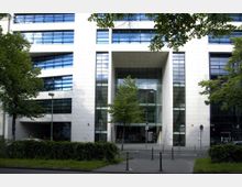 Moderne Bürogebäude-Fassade mit großen Fensterfronten, weißem Stein und einem überdachten Eingangsbereich. Im Vordergrund befinden sich Gehwege, Poller, ein schmaler Grünstreifen sowie Bäume, die Teile der Fassade verdecken.