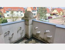 Balkon mit gealtertem Putz und Verzierungen an der Brüstung, bestehend aus durchbrochenen Steinornamenten; im Hintergrund Wohnhäuser mit roten Dächern und Bäumen in einer ruhigen Nachbarschaft.