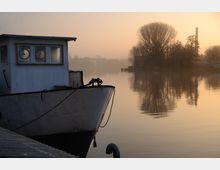 Ein stillgelegtes Boot liegt an einem Steg in ruhigem Wasser, umgeben von einer nebligen Morgenstimmung. Im Hintergrund sind silhouettenartige Bäume und ein Schornstein im sanften orangefarbenen Licht der aufgehenden Sonne sichtbar.