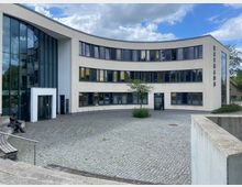 Moderne Rathausgebäude mit geschwungener, heller Fassade und großen Fenstern. Der Vorplatz besteht aus einem mit Pflastersteinen belegten Bereich, auf dem ein Baum und eine Sitzbank mit einer Bronzestatue zu sehen sind.