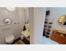 Kleines Badezimmer mit weißen Wandfliesen, einem hängenden WC, einem Waschbecken und einem runden Spiegel. Rechts daneben befindet sich ein Flur mit weißen Holzfußböden, einem Schuhregal, einem Holzschrank und einem weiteren runden Spiegel.