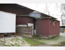 Blechverkleidetes Gebäude mit brauner Fassade und rostfreiem Metalltank auf dem Dach, daneben ein weißer Anhänger. Der Boden ist teils mit Betonplatten, teils mit Gras und Sand bedeckt.