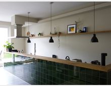 Moderne Küche mit einem langen Tresen, der mit glänzenden, grünen Fliesen verkleidet ist und einer Holzarbeitsplatte. Über dem Tresen hängen schwarze Pendelleuchten, an der Rückwand befindet sich ein schlichter Holzregal mit Dekorationen, und eine große Dunstabzugshaube ist sichtbar.