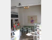 Ein kleines Café mit heller Einrichtung, weißen Wänden und einer verspielten Dekoration. Es gibt weiße Tische und Stühle, eine Theke mit Gebäck, eine gläserne Vitrine mit Karten sowie Wandbilder, darunter ein großes Gemälde eines Kindes.