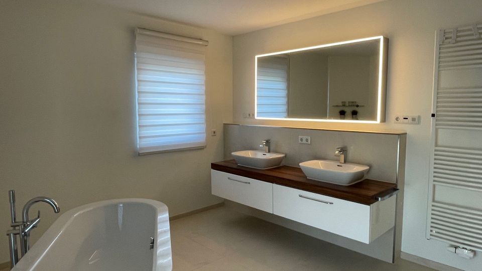 Helles modernes Badezimmer mit freistehender Badewanne, Doppelwaschtisch mit Holzplatte und rechteckigem Spiegel mit umlaufender Beleuchtung. An der Wand befinden sich ein Heizkörper und ein Fenster mit gefaltetem Rollostoff.