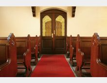 Innenraum einer Kirche mit hölzernen Kirchenbänken auf beiden Seiten eines roten Teppichgangs, der zu einer dunkelbraunen, verglasten Tür mit verziertem Gittermuster führt. Die Wände sind schlicht hell gehalten, und die Türrahmen sind mit Holzdetails versehen.