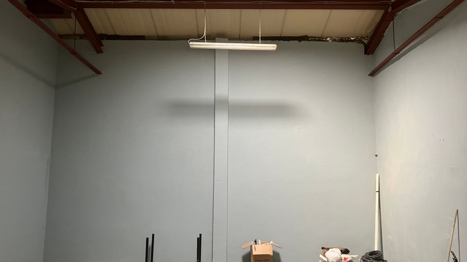 Empty warehouse-style interior with tall gray walls, exposed red steel ceiling beams, and a hanging fluorescent light fixture. Concrete floor area meets a section of wood-pattern flooring, with a few boxes and coiled cables stacked along one side.