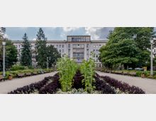 Ein gepflasterter Parkweg führt zu einem großen, mehrstöckigen Gebäude mit heller Fassade und symmetrischen Fenstern im Hintergrund. Der Weg ist von Blumenbeeten mit bunten Pflanzen und Sitzbänken gesäumt, flankiert von hohen Bäumen und Laternen.