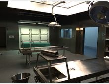 Ein steriler Raum in einer Pathologie mit Edelstahl-Arbeitsflächen, Obduktionstischen und Deckenleuchten. Im Hintergrund sind Schrankfächer mit Türen und ein Fenster zu sehen; die Wände sind dunkel und der Boden grau.