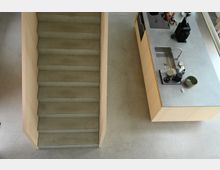 Treppenaufgang und Küchentresen in einer modernen, minimalistischen Innenraumgestaltung mit Betonboden, Holzgeländer und einer Arbeitsfläche mit Spüle und Küchengeräten.