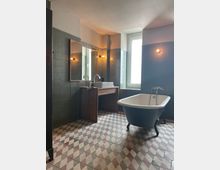 Ein Badezimmer mit einer freistehenden weißen Badewanne auf schwarzen Beinen, einem Holzwaschtisch mit Aufsatzwaschbecken und einem großen Spiegel an der Wand. Die Wände sind mit grünen Fliesen verkleidet, der Boden ist mit gemusterten Fliesen in Rautenmuster gestaltet, und ein großes Fenster lässt Tageslicht herein.