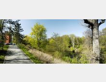 Weg in einer Parklandschaft mit Bäumen und Büschen, die teilweise frisches Frühlingsgrün zeigen. Rechts befindet sich ein kleiner Teich, links parken einige Fahrzeuge vor einem roten Backsteingebäude.