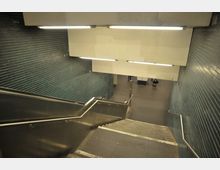 Treppenabgang in einer U-Bahn-Station mit grünen Fliesenwänden, metallenen Handläufen und heller Deckenbeleuchtung. Unten führt die Treppe in einen offenen Bereich mit grauem Bodenbelag.