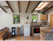 Kleine Wohnküche mit rustikalem Stil: Sichtbare Holzbalken an der Decke, Fliesenboden, eine Küchenzeile mit gemusterten Fliesenspiegel und einem Backofen, sowie ein Kühlschrank. Zwei Fenster bieten Blick ins Grüne, und im Raum stehen eine Holzbank, ein kleiner Beistelltisch, eine Standuhr und Dekorationen.