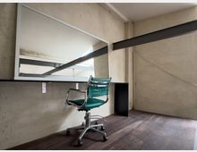 Ein minimalistischer, moderner Raum mit einem großen rechteckigen Wandspiegel, einem schlichten Holztisch und einem grünen Bürostuhl auf Rollen. Der Raum hat beige Wände, sichtbare Metallträger und einen dunklen Holzboden.