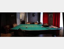 Ein historisch eingerichteter Raum mit einem großen Roulette-Tisch in der Mitte, auf dem Chips und eine Roulettekessel liegen. Im Hintergrund befinden sich rote Vorhänge, ein Polstersessel, eine geöffnete Tür und dekorative Tapeten an den Wänden.