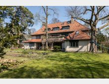 Großes Wohnhaus mit rotem Satteldach und mehreren Dachgauben, umgeben von einem gepflegten Garten mit Bäumen und Rasenfläche. Im Vordergrund ist eine offene Grünfläche, im Hintergrund vereinzelte Gebäude und Bäume.