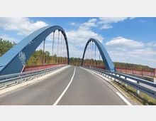 Moderne Straßenbrücke mit zwei blauen Stahlbögen und roten Streben über einem Fluss, umgeben von Wald und klarem Himmel mit einigen Wolken. Die Fahrbahn ist zweispurig und von Leitplanken begrenzt.