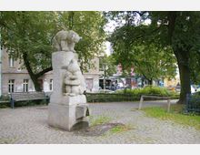 Steinskulptur mit Tierfiguren, darunter ein Bär und ein Tier mit rundlichen Formen, auf einem Platz mit Pflastersteinen und umgeben von Bäumen. Im Hintergrund sind Bänke, ein Gebäude mit Graffiti und eine Straße mit parkenden Autos zu sehen.