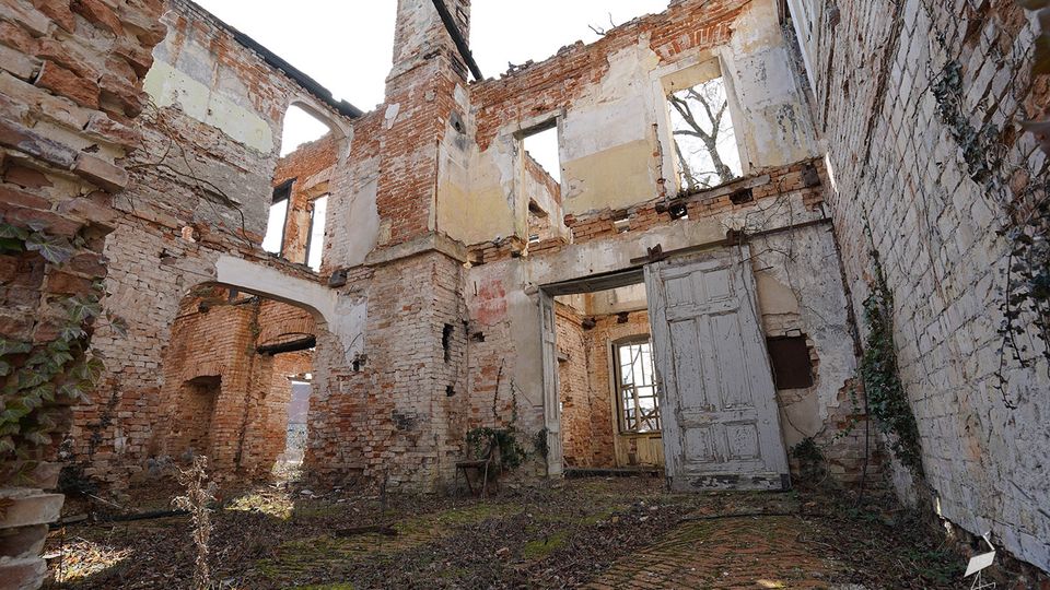 Innenhof eines verfallenen Backsteingebäudes ohne Dach, mit freiliegenden Ziegelwänden, großen leeren Fensteröffnungen und einem gemauerten Bogen. Im Vordergrund liegen überwucherte Bodenflächen und eine verwitterte Tür in einem teilweise erhaltenen Innenraum.