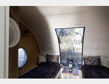 Innenraum einer kleinen, hölzernen Unterkunft mit gewölbter Decke. Zwei Einzelbetten mit gemusterten Kissen stehen nebeneinander, mit einer großen, schrägen Fensterfront und Blick auf Schilf und eine Terrasse; ein rundes Seitenfenster ergänzt die Einrichtung.