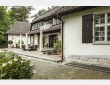 Einfamilienhaus mit weißer Fassade, einem dunklen, steilen Ziegeldach und Sprossenfenstern, umgeben von Bäumen. Im Vordergrund befindet sich eine Terrasse mit Holzmöbeln, Sonnenschirmen und Blumen in Töpfen.