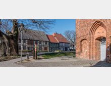 Fachwerkhäuser mit dunklen Holzrahmen und roten Dächern stehen an einem gepflasterten Weg, neben einer großen Backsteinkirche mit Spitzbogenportal und weißer Tür. Im Vordergrund sind ein alter Baum und eine Laterne zu sehen, die Szene spielt an einem sonnigen Tag.