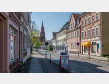Straßenszene in einer kleinen Stadt mit gepflasterter Fahrbahn, Bürgersteig und aneinandergereihten Wohn- und Geschäftshäusern. Im Hintergrund steht eine rote Backsteinkirche mit hohem Turm, und Bäume säumen den Straßenrand.