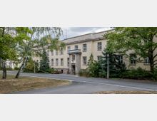 Zweistöckiges Gebäude mit heller Fassade und symmetrischen Fenstern, umgeben von Bäumen und Büschen. Im Vordergrund führen gepflasterte Wege an einer kleinen Grünfläche mit Bänken und Straßenlaternen vorbei.