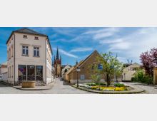 Straßenszene in einer Altstadt mit Kopfsteinpflaster, flankiert von einem hellen Wohngebäude links und einem gelblichen Haus mit Satteldach rechts. Im Hintergrund ragt eine Kirche mit rotem Mauerwerk und mehreren spitzen Türmen empor, während im Vordergrund Blumenbeete und ein Baum einen kleinen Platz umgeben.
