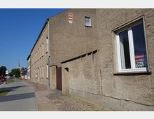Straßenansicht mit einem länglichen, beige verputzten Wohngebäude und einem schmalen Bürgersteig; im Hintergrund ragt der Turm einer Kirche hervor, daneben weitere Gebäude. Ein Fenster trägt ein Schild mit der Aufschrift "Wohnung zu vermieten".