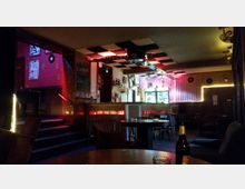 Ein gemütlicher Barraum mit gedämpfter Beleuchtung, Holztischen und Stühlen. Im Hintergrund befindet sich eine Theke mit Flaschenregalen und beleuchteten Kühlschränken, die von dekorativen Lichtern sowie Wandverkleidungen mit Platten und Postern umgeben ist.