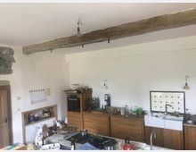 Rustikale Küche mit weiß verputzten Wänden und einer sichtbaren Holzbalkendecke. Die Einrichtung besteht aus hölzernen Schränken, einer großen weißen Spüle mit Fliesenspiegel und offen sichtbaren Küchenutensilien.