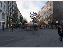 Fußgängerzone in einer Stadt mit modernen Geschäftsgebäuden auf beiden Seiten, Geschäften, einer Apotheke und Sitzbereichen unter Sonnenschirmen in der Mitte. Laternen und eine gepflasterte Fläche prägen den Platz, Passanten beleben die Szene.