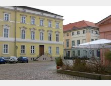 Kleiner gepflasterter Platz, umgeben von historischen Gebäuden mit gelben und pastellfarbenen Fassaden, verziert mit Stuckdetails. Im Vordergrund geparkte Autos, Laternen und ein weißes Zelt; einige Pflanzen säumen den Bereich.