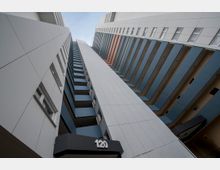 Hochwinkelansicht eines modernen, mehrstöckigen Wohn- oder Bürogebäudes mit weißen Fassaden und blauen Balkonverkleidungen. Die Nummer „120“ ist über einem Eingangsbereich sichtbar, und die Perspektive betont die vertikale Höhe der Struktur.