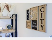 Ein dekoratives Innenraumdetail mit einem Holzregal, das getrocknete Gräser in Vasen, eine Uhr und Kerzen enthält, sowie einer an der Wand hängenden Holzwanddekoration mit Schriftzügen wie „Keep Smile“, „& Be Happy“ und einer stilisierten Fahrradzeichnung.