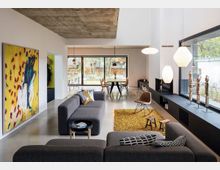 Modernes Wohnzimmer mit grauen Sofas, einem gelben Teppich und Wandkunst auf der linken Seite. Große Fensterfronten lassen viel Tageslicht herein und bieten Blick auf einen Garten, während hängende Lampen und eine Sichtbetondecke das Design abrunden.