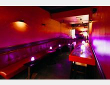 Schmaler Lounge- oder Barbereich mit roten Wänden und durchgehender, gepolsterter Sitzbank entlang der linken Seite. Kleine Tische mit violett leuchtenden Tischlampen und ein langer Hochtisch rechts führen zu einem Spiegel am Ende des Raums.