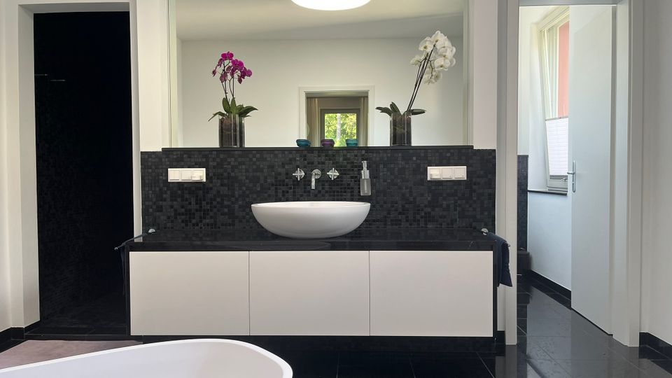 Modernes Badezimmer mit schwarzem Fliesenboden und schwarzer Mosaikrückwand, davor ein schwebender weißer Waschtischunterschrank mit ovalem Aufsatzwaschbecken und großer Spiegelwand. Im Vordergrund ist der Rand einer freistehenden Badewanne zu sehen, rechts führt eine Tür in einen hellen Nebenraum mit Fenster.