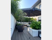 Moderne Terrasse mit Holzfußboden, umgeben von weißen Wänden und dichter Bepflanzung in großen Töpfen. Im Mittelpunkt steht ein schwarzer Sonnenschirm über einer Sitzecke mit hellen Polstermöbeln, ergänzt durch Pflanzen in Blumenbeeten und Töpfen.